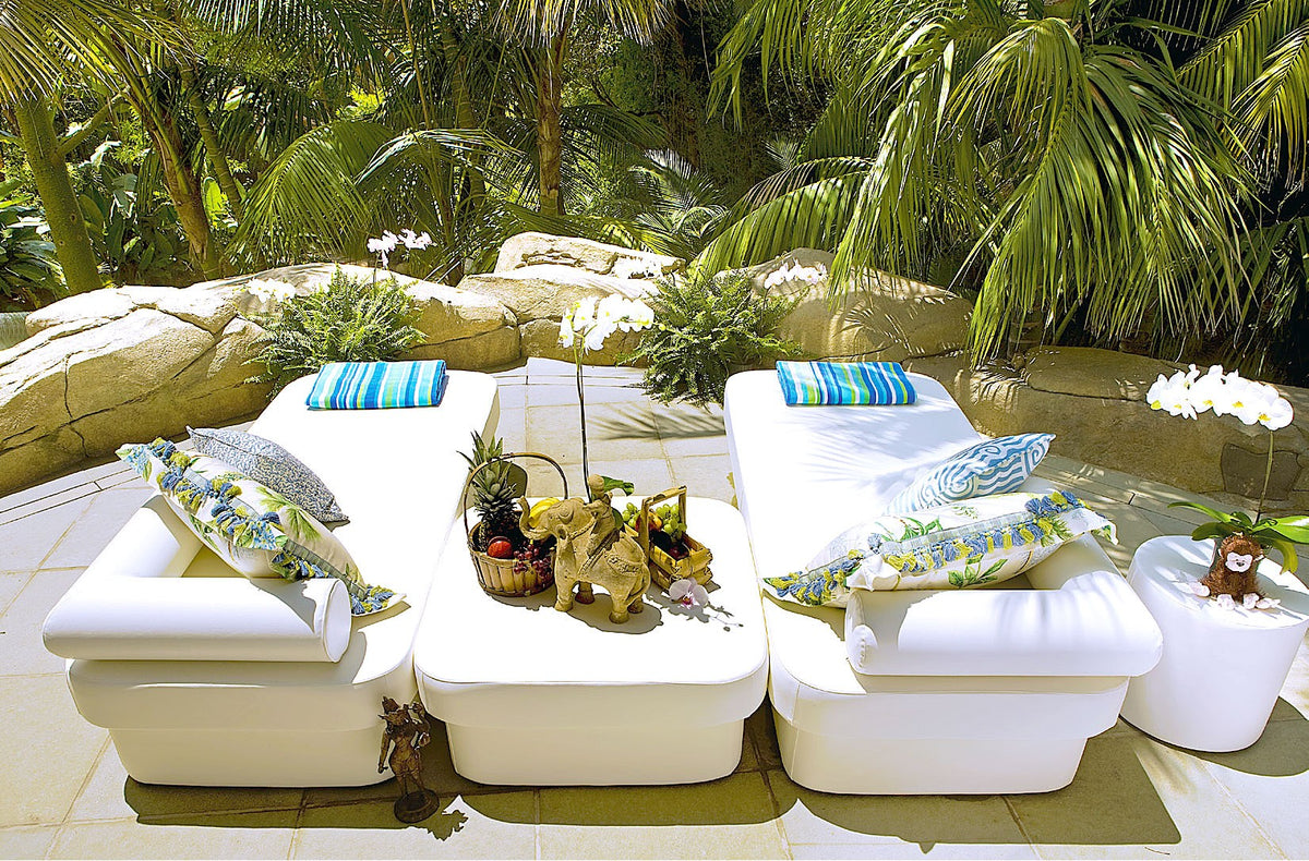 Pair of white Cot twin daybeds with striped pillows and a fruit basket beside a garden pond with palms