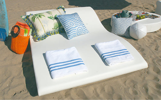 Wide white La Fête Tide double lounge on a beach with two folded striped towels and patterned pillows