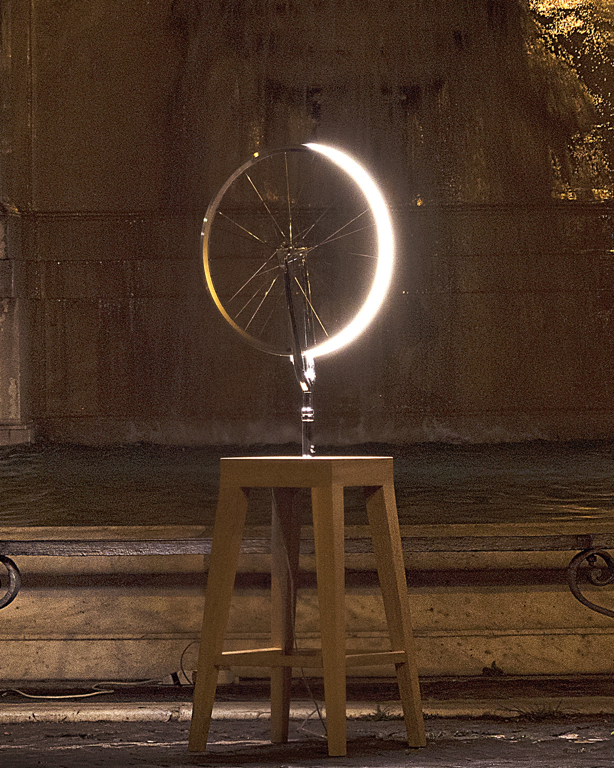 Cyclampa Marcel lamp lit at night, glowing wheel rim on oak stool